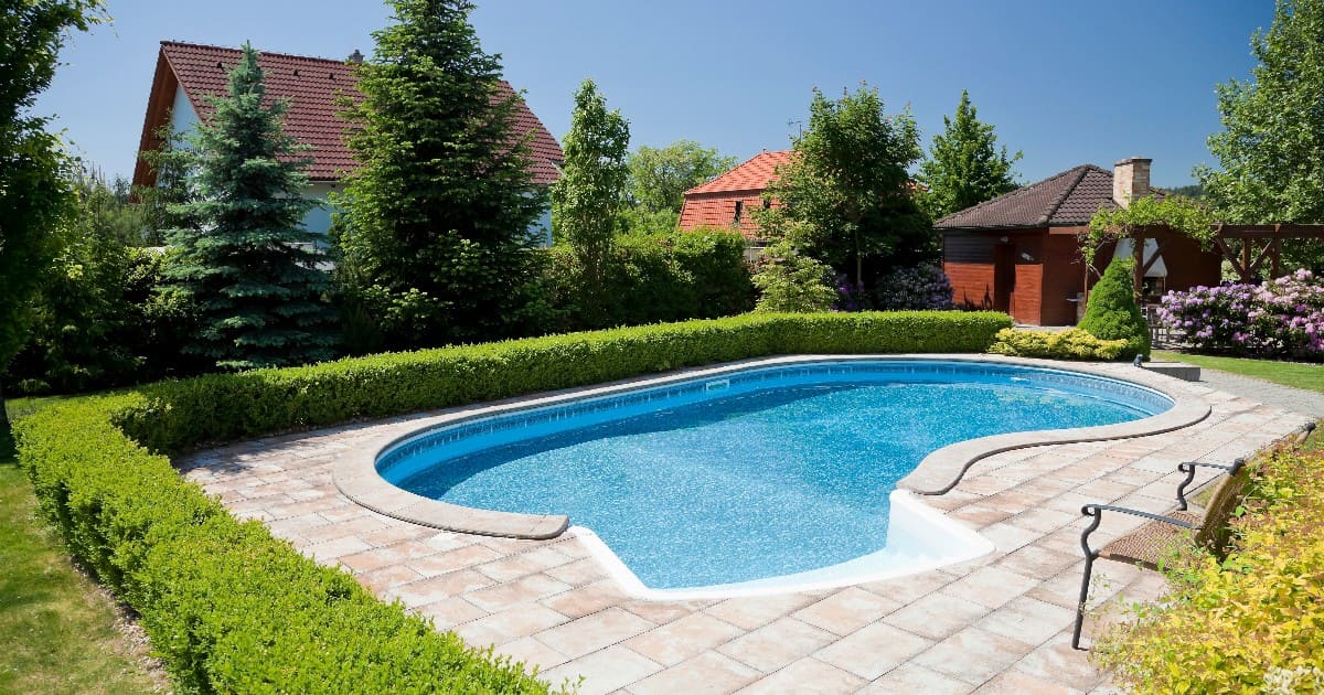A curved inground swimming pool surrounded by manicured green hedges and a stone patio on a sunny day, showcasing a landscaping project when considering how long does it take to build a pool.