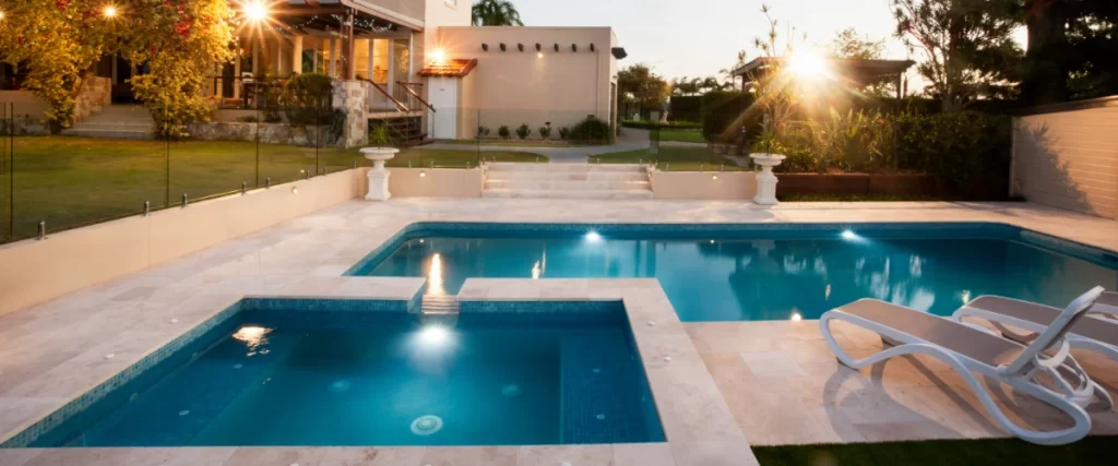 A rectangular backyard pool beautifully illuminated at sunset, featuring a square integrated spa, a shallow tanning ledge, and surrounded by light-colored stone pavers.