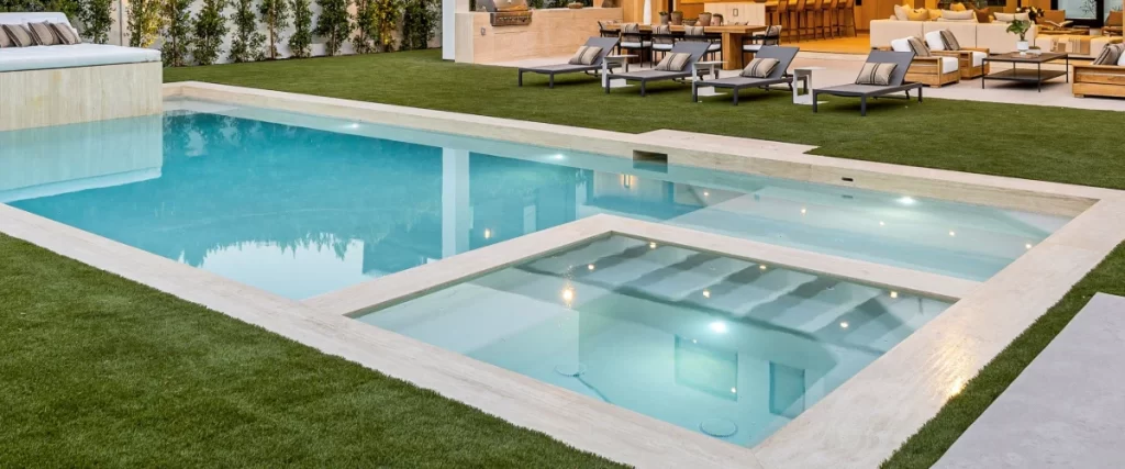 A modern geometric swimming pool surrounded by artificial grass, showcasing a built-in square spa and a wide, shallow tanning ledge with submerged entry steps.