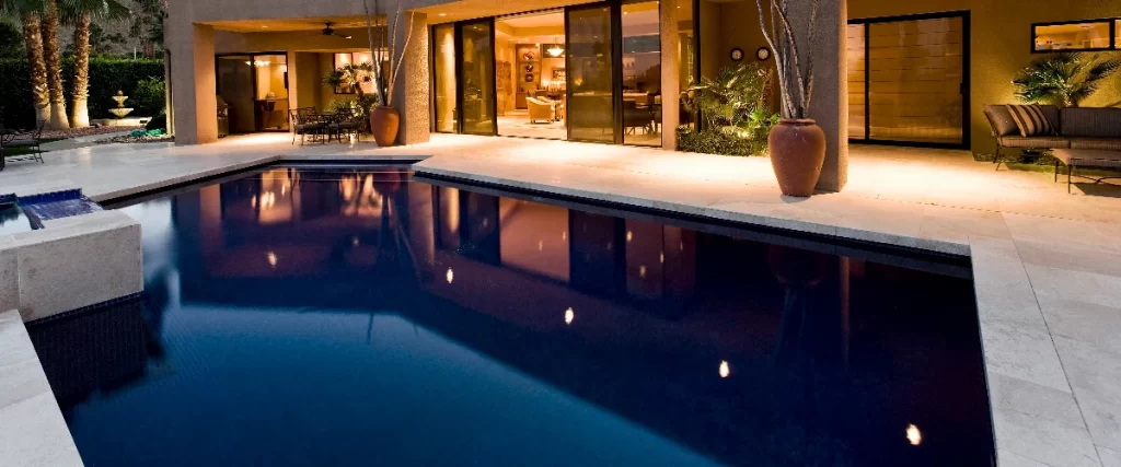 A modern, dark-bottomed rectangular pool reflecting the warm evening lights of a luxurious outdoor patio and open glass doors.