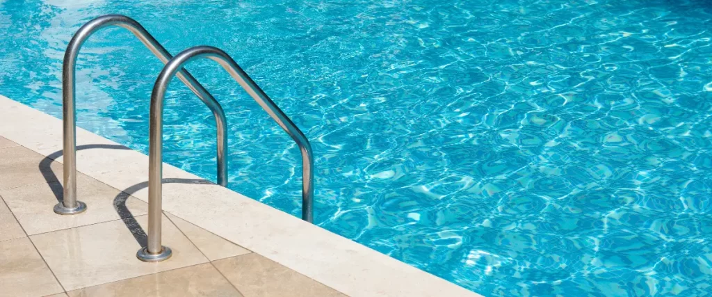 A close-up view of polished stainless steel ladder handrails securely mounted to stone coping, leading into bright blue, rippling pool water.