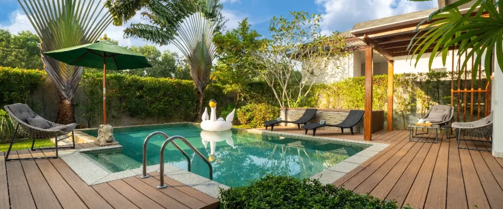 A tropical backyard swimming pool featuring a wooden deck, comfortable lounge chairs under a green umbrella, and a large white swan float resting on the water.
